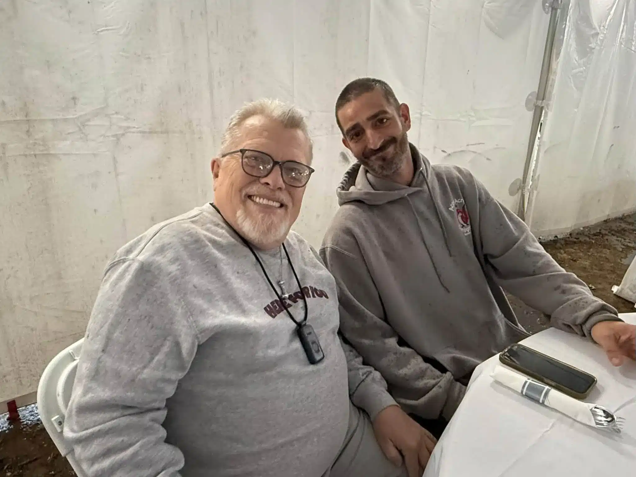 Two smiling men seated side by side against a white event tent wall at Acorn Acres Campground in Bozrah, CT